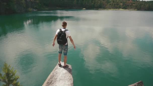 Jovem homem ativo alarga as mãos na encosta da rocha sobre a superfície calma lago montanha — Vídeo de Stock