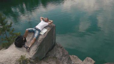 Onun yanında döşeme harita ile sakin dağ gölü yüzeye yakın kaya üzerinde bekleterek genç turist adam rahatlatır