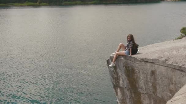 Joven mochilero atractiva chica se relaja descansando en la roca de montaña sobre la superficie del agua del lago por debajo — Vídeos de Stock