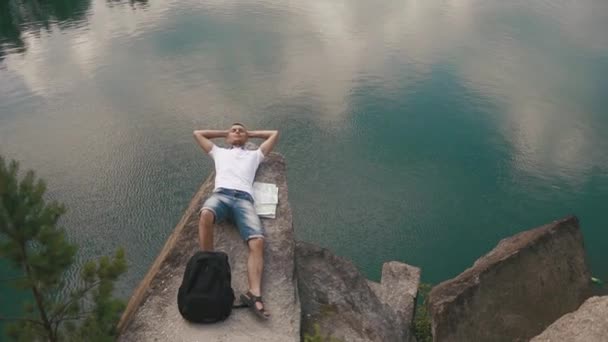 Jovem homem ativo relaxa descansando na encosta da rocha perto da superfície calma lago montanha com mapa que coloca ao lado dele — Vídeo de Stock