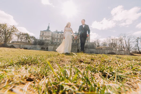 Atrakcyjne nowożeńcy para chodzenie na zielonym słonecznym trawniku w pobliżu pięknego zrujnowanego barokowy pałac. Kochający Pan młody trzyma uroczą narzeczoną ręką — Zdjęcie stockowe