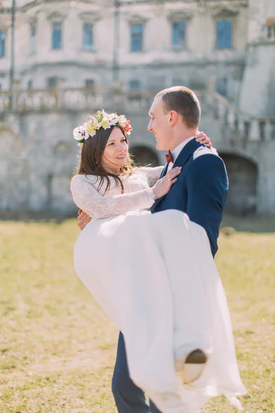 Atraente casal recém-casado no gramado verde perto de belo palácio barroco arruinado. Noivo amoroso segurando noiva encantadora em suas mãos — Fotografia de Stock