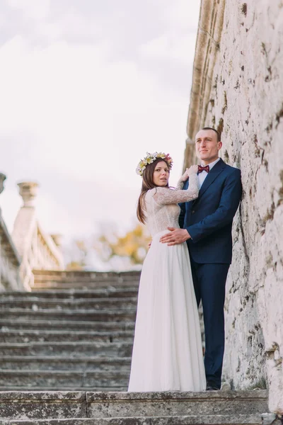 Feliz novio elegante sosteniendo a su novia bonita, mientras que ambos de pie en escaleras de piedra antigua. Retrato de cuerpo entero —  Fotos de Stock