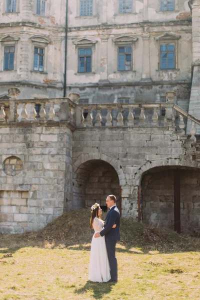 Charming newlywed bride and groom holding each other on lawn near beautiful ruined baroque palace — Stock Photo, Image