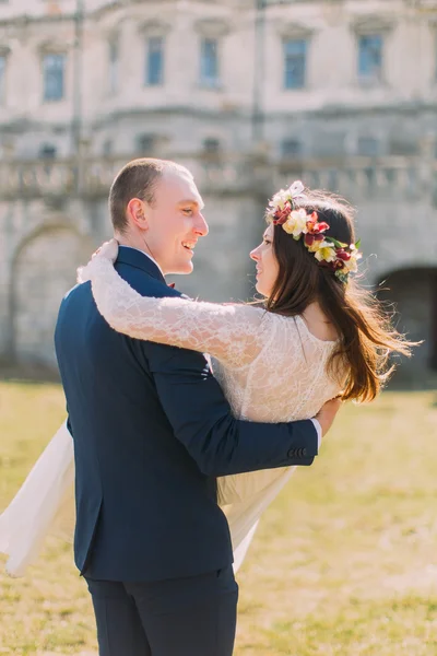 Attractive newlywed pair at green sunny lawn near beautiful ruined baroque palace. Loving groom holding charming bride on his hands — Stock Photo, Image