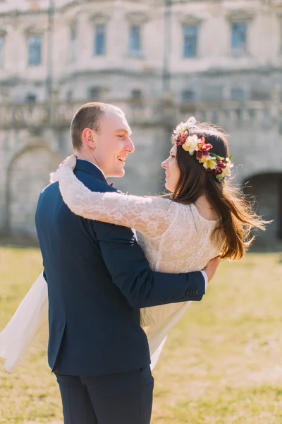 Attraente coppia di sposi appena sposati a prato verde e soleggiato vicino al bellissimo palazzo barocco in rovina. Amare sposo tenendo sposa affascinante sulle sue mani — Foto Stock