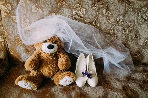Кукла плюшевого мишки вместе с свадебными аксессуарами - обувью и вуалью - изолированы на диване. Чувственная свадебная концепция — стоковое фото