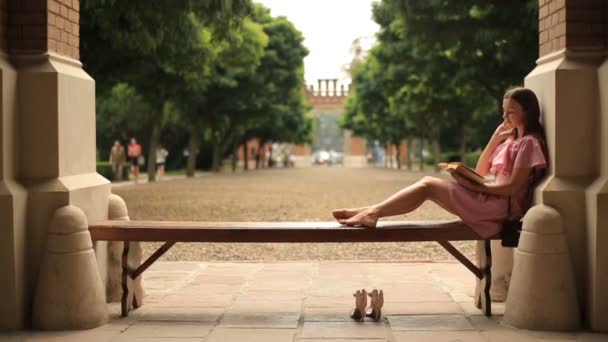 Красивая девочка-подросток лежит на скамейке запасных в университетском городке и читает книгу босиком — стоковое видео