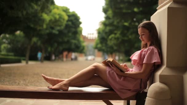 Adolescente sentada en el banco en la naturaleza y leyendo un libro — Vídeos de Stock