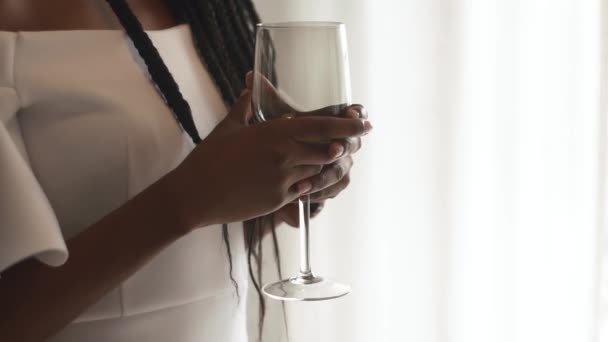 Joven modelo femenina afroamericana sosteniendo en su mano un vaso de vino vacío, de pie cerca de una ventana iluminada con cortinas blancas — Vídeos de Stock