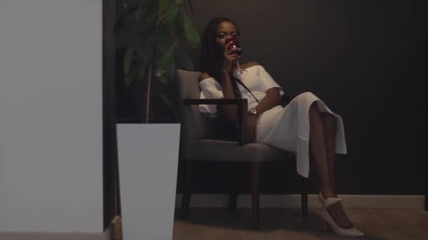 Young beautiful african american female fashion model drinking from red wineglass while sitting in comfortable chair — Stock Video