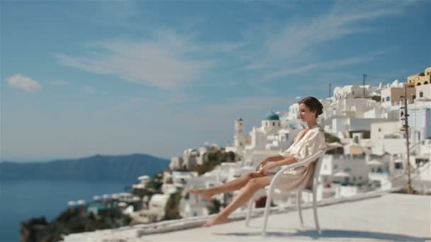 Linda noiva morena sexy em roupão sentado na cadeira branca e acenando com as pernas desfrutando da vista do mar e das montanhas — Vídeo de Stock