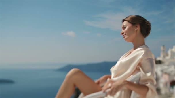 Belle mariée brune sexy en robe assise sur la chaise blanche enjoyng la lumière du soleil et une vue imprenable sur la mer et les montagnes en arrière-plan — Video