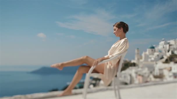 Belle mariée brune sexy en robe assise sur la chaise blanche serrant ses jambes et enjoyng la vue sur la mer et les montagnes en arrière-plan — Video