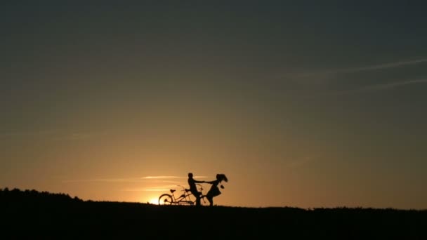 すばらしい夕焼け背景に対して手を繋いで踊るタンデム自転車で美しいカップルのシルエット . — ストック動画