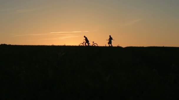 Silueta de hermosa pareja con bicicletas en tándem. El hombre monta la bicicleta en tándem mientras su novia lo atrapa con gracia contra la puesta del sol. Nubes de verano en el cielo de cerca — Vídeo de stock