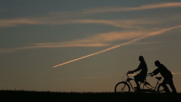 美丽的夫妇与双座自行车的轮廓。女孩骑着自行车，虽然他推它夏天自然背景与美丽的夕阳和天空中的云彩关闭 — 图库视频影像