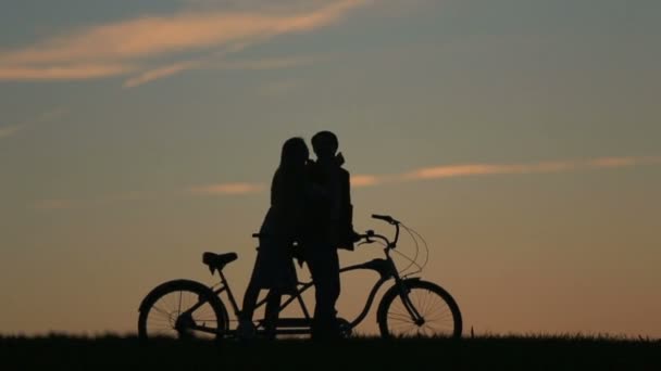 Silueta de hermosa pareja con bicicletas en tándem. Chica acaricia sus mejillas. Fondo de la naturaleza de verano con hermosa puesta de sol y nubes en el cielo de cerca . — Vídeos de Stock