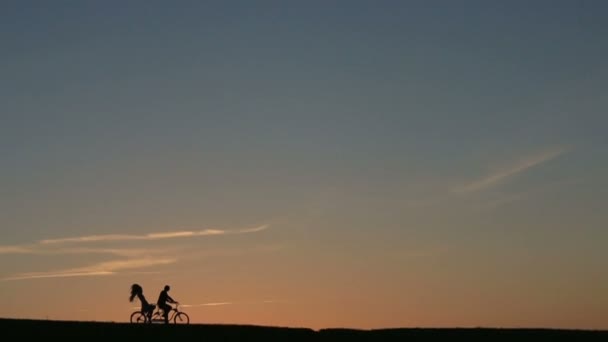 Sziluettje gyönyörű pár lovaglás a Tandem kerékpárok, míg a naplemente. Nyári természet háttér gyönyörű felhők az égen — Stock videók