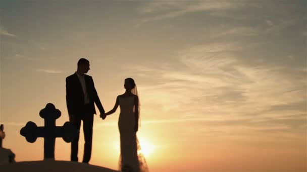 Feliz pareja casada caminando tomados de la mano en el techo de la iglesia al atardecer cielo fondo — Vídeo de stock