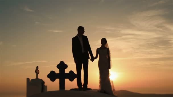 Feliz pareja casada caminando de la mano y besándose en el techo de la iglesia al atardecer fondo del cielo — Vídeos de Stock
