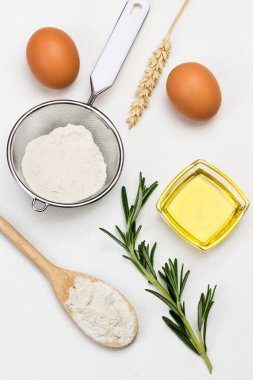 Tahta kaşıkta un. Zeytinyağı ve biberiye filizi, elek ve cam kavanoz. Yumurta ve dikenli buğday. Beyaz arka plan. Üst görünüm