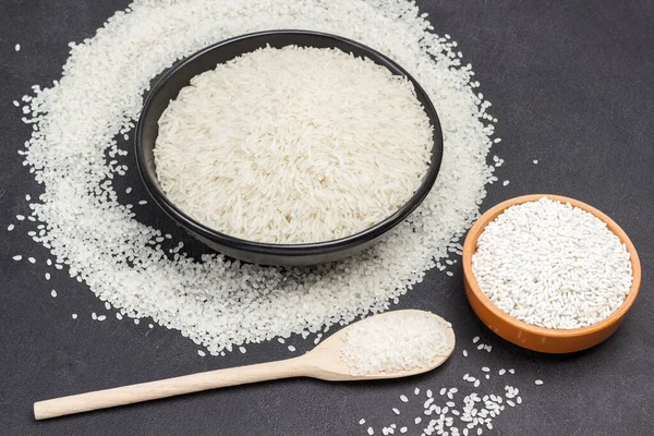 Basmati rice in black bowl. Glutinous rice in ceramic bowl. White round rice in wooden spoon and scattered on table. Black background. Top view