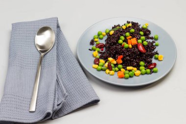 Gri tabakta sebzeli siyah pirinç. Kaşık gri peçeteye. Gri arka plan. Üst görünüm