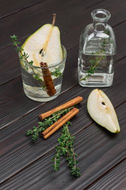 Armut suyu, tarçın çubuğu ve kekik şeritli cam. Masada bir dilim armut. İçinde su ve kekik filizi olan bir şişe. Karanlık ahşap arka plan. Üst görünüm
