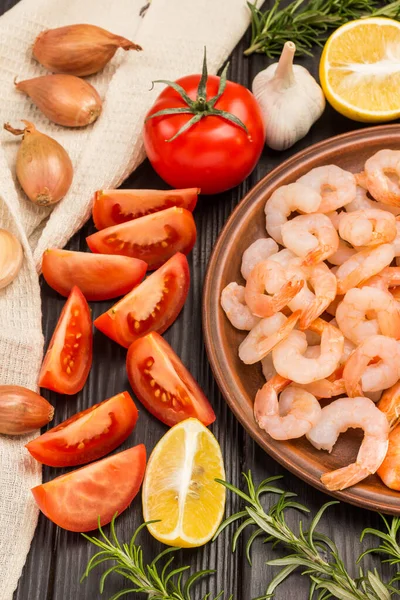 Shrimp in ceramic plate. Tomatoes, lemon, rosemary sprigs on table. Onions on beige napkin. Dark wooden background. Top view