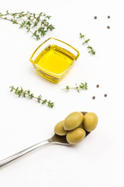 Metal kaşıkta yeşil zeytin. Cam kavanozda zeytinyağı. Kekik filizleri. Beyaz arka plan. Üst görünüm