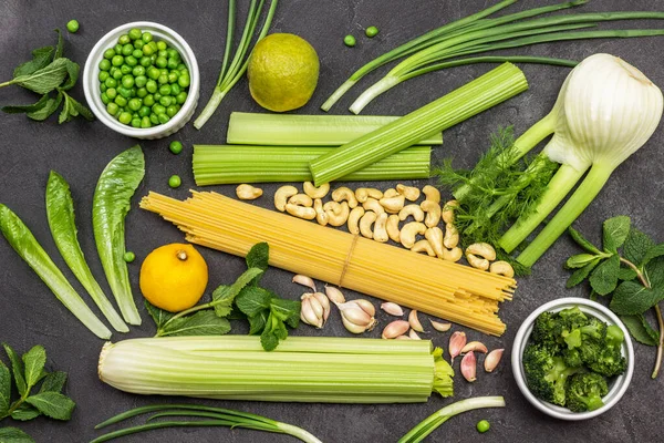 Set of vegetables and pasta. Celery stems, fennel and mint. Cashew nuts and chives. Broccoli and green peas in ceramic bowls. Black background. Flat lay