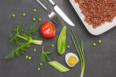 Yumurta, domates ve otların yarısı masada. Beyaz tabakta esmer pirinç. Çatal ve bıçak masada. Dengeli diyet. Siyah arka plan. Düz yatıyordu