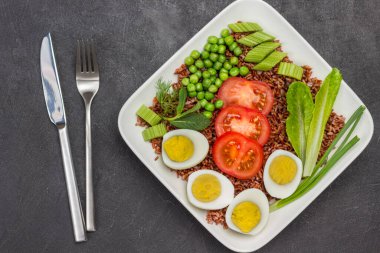 Kahverengi pirinç, yumurta ve sebzeler beyaz tabakta. Çatal ve bıçak masada. Dengeli yemek. Siyah arka plan. Üst görünüm