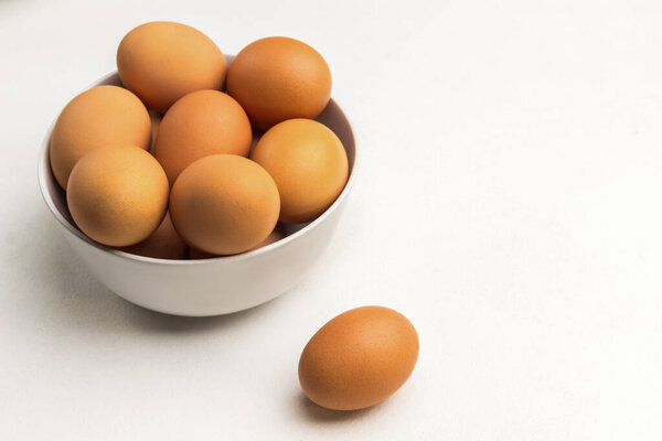 Brown eggs in white ceramic bowl. One egg on table. Copy space. White background. Top view