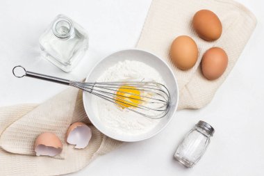 Kırık yumurta ve çatal gri kasede. Kağıt üzerinde un ve elek. Gri tabakta tavuk kabukları. Kahverengi yumurtalar masada. Gri arkaplan. 