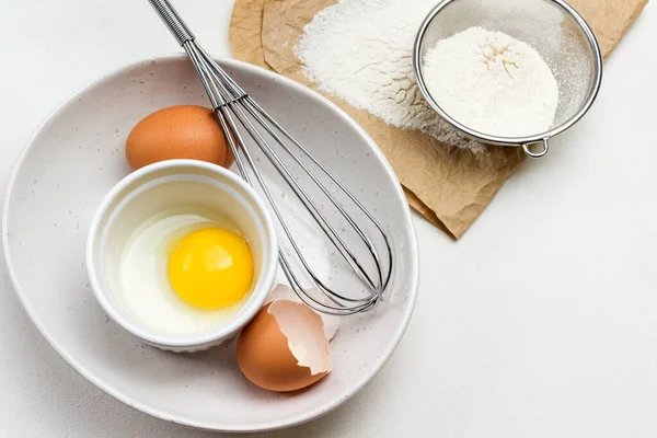 Yumurta sarısı küçük kasede. Tavuk yumurtası, yumurta kabuğu ve büyük beyaz kasede çırpma. Kağıt üzerinde un ve elek. Beyaz arka plan. Düz yatıyordu