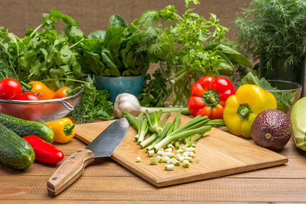 Sliced green onions, kitchen knife on cutting board. Vegetables and dill, spinach leaves and cilantro on table. Wooden background