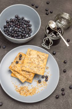 Gri tabağa tohum serpiştirilmiş bisküviler. Reçel kavanozu. Gri kasede yabanmersini. Siyah arka plan. Düz yatıyordu