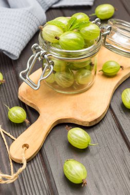 Kesme tahtasındaki cam kavanozda yeşil bektaşi üzümü. Masada gri peçete ve bektaşi üzümü var. Koyu ahşap arka plan. Üst görünüm