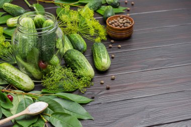 Fresh cucumbers in glass jar. Cucumbers, dill and cherry sprig on table. Salt in wooden spoon.  Copy space. Wooden background. Top view