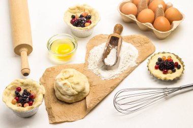 Kağıt üzerinde un ve hamur. Donmuş böğürtlen ve hamur dolu fırın kabı. Çırpı, yumurta tepsisi ve yumurta sarısı kasede. Yumurta tepsisi