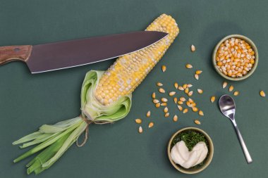 Çiğ mısırın üzerinde bıçak. Masada ve kasede mısır taneleri var. Kasede sos. Gri arka plan. Düz yatıyordu