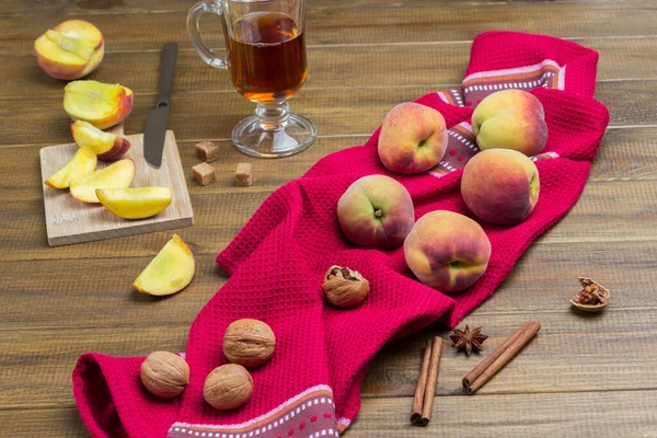 Peaches and walnuts on red napkin. Sliced peaches and knife on board. Glass of tea, cinnamon sticks and star anise on table