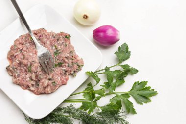 Kıyma ve çatal tabakta. Masada soyulmuş soğan ve maydanoz var. Beyaz arka plan. Üst görünüm