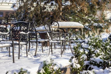 Kışın bir kafe terası, karla kaplı masalar ve sandalyeler. Yerde kar ve ağaçlar.
