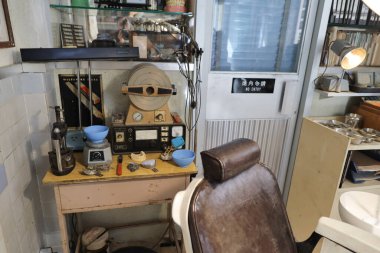 A retro dental room features a worn brown chair, overhead lamp, and glass shelves