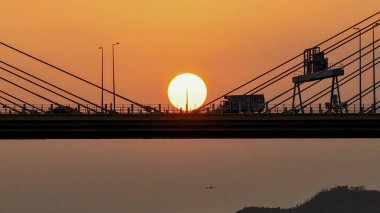 Köprü siluetleri, sakin suyun üzerinde parlayan bir günbatımını çerçeveler.