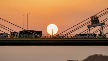 Köprü siluetleri, sakin suyun üzerinde parlayan bir günbatımını çerçeveler.