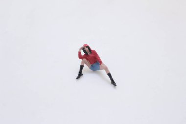 A tiny plastic figurine sits on a clean white surface, wearing a red hoodie, b